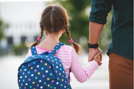A girl with pigtails, wearing a polka-dotted backpack, holds hands with an adult.