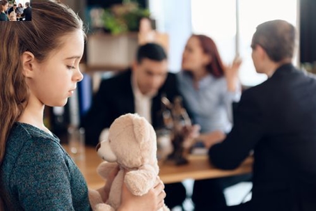 A sad young girl clutches a teddy bear, while in the background two adults argue