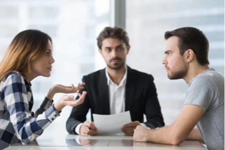 A couple sitting at a desk during a tense discussion with a mediator or lawyer holding a document.