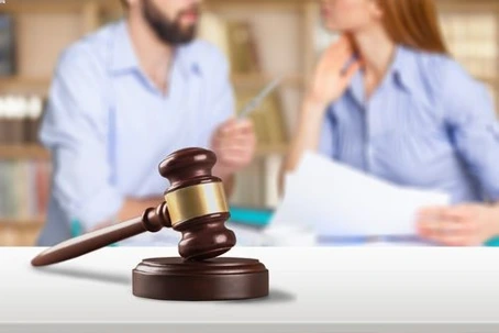 A wooden judge's gavel rests on a surface in the foreground, with a blurry background showing a man and a woman discussing documents