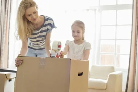 A woman and a young girl packing and taping cardboard boxes together.