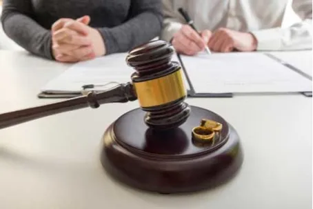 A judge's gavel and wedding rings on a sounding block with people signing papers in the background.