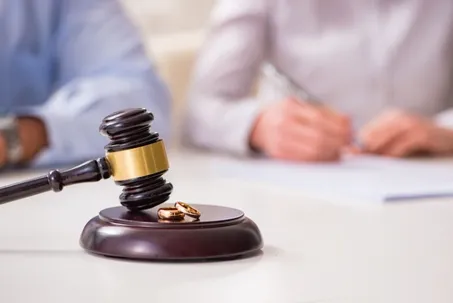 A judge's gavel and two wedding rings resting on a block, with people signing documents in the background.
