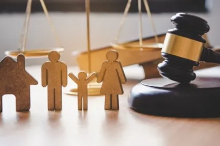 A wooden house figure, family figures, a set of scales, and a judge's gavel on a table.
