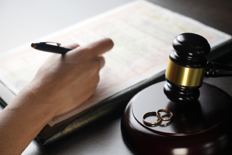 Close-up of hand signing divorce or marriage papers with pen, next to judge gavel and two golden wedding rings. Concept of divorce, family law, marriage, legal separation, court decision and justice